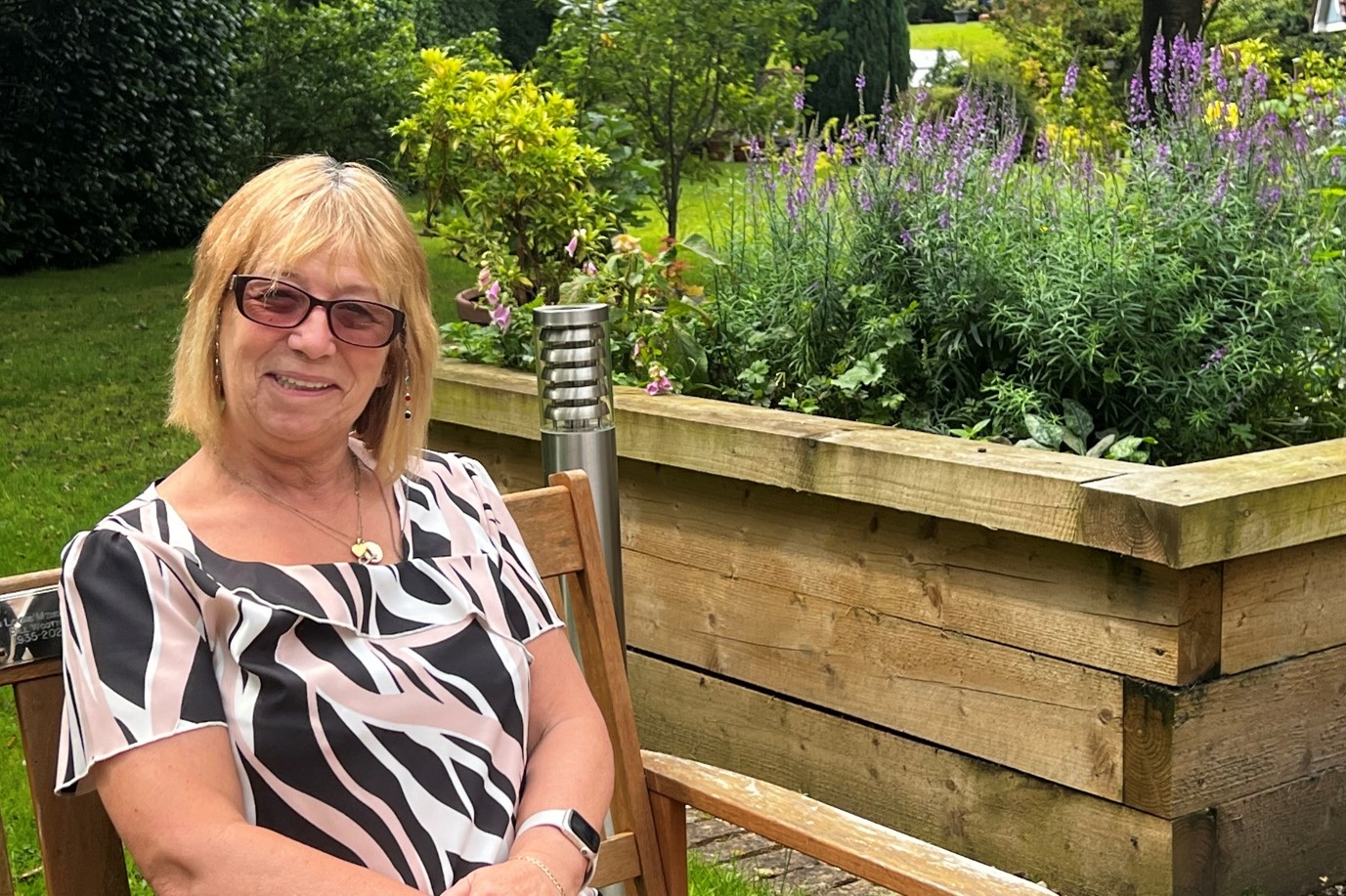 Older woman sat in her garden looking at the camera and smiling.