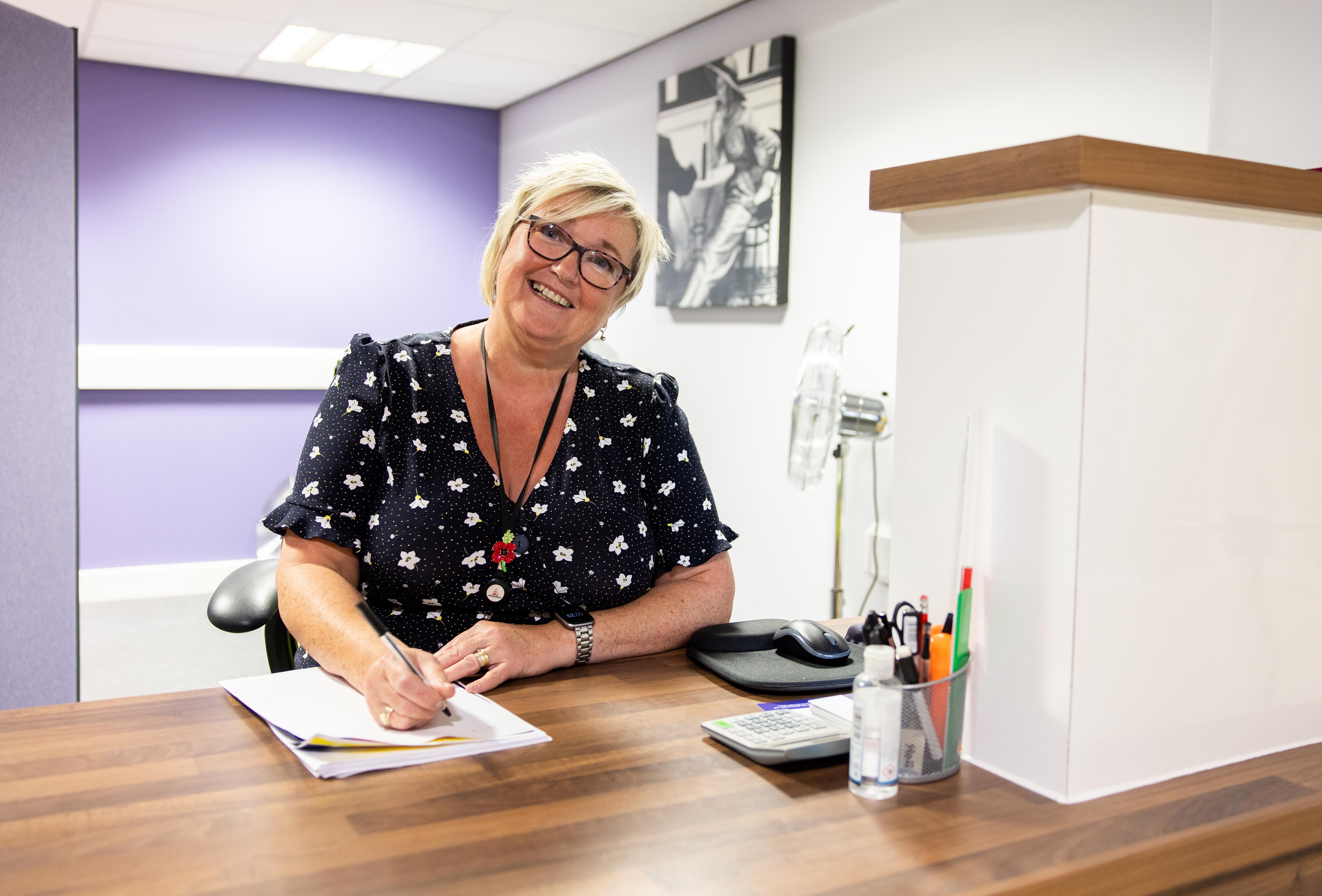 Woman sat at a reception desk, looking at the camera and smiling