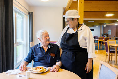 Older man sat having his breakfast, with a kitchen staff member talking to the man, with their arm around them, both are looking at each other and laughing and smiling. 