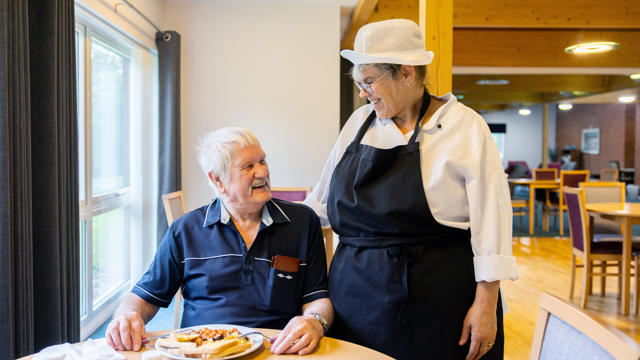 Older man sat having his breakfast, with a kitchen staff member talking to the man, with their arm around them, both are looking at each other and laughing and smiling. 