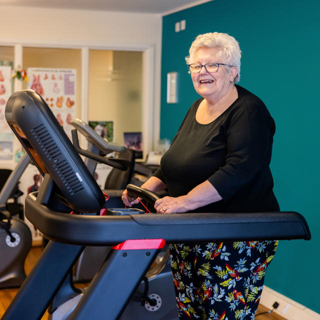 Older woman stood on a treadmill, looking at the camera and smiling. 