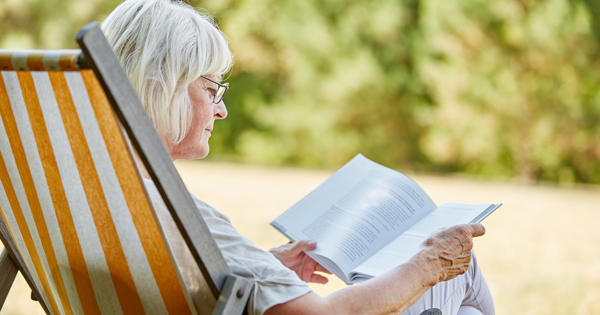 Older Woman Reading A Book In The Garden On A Deck Chair